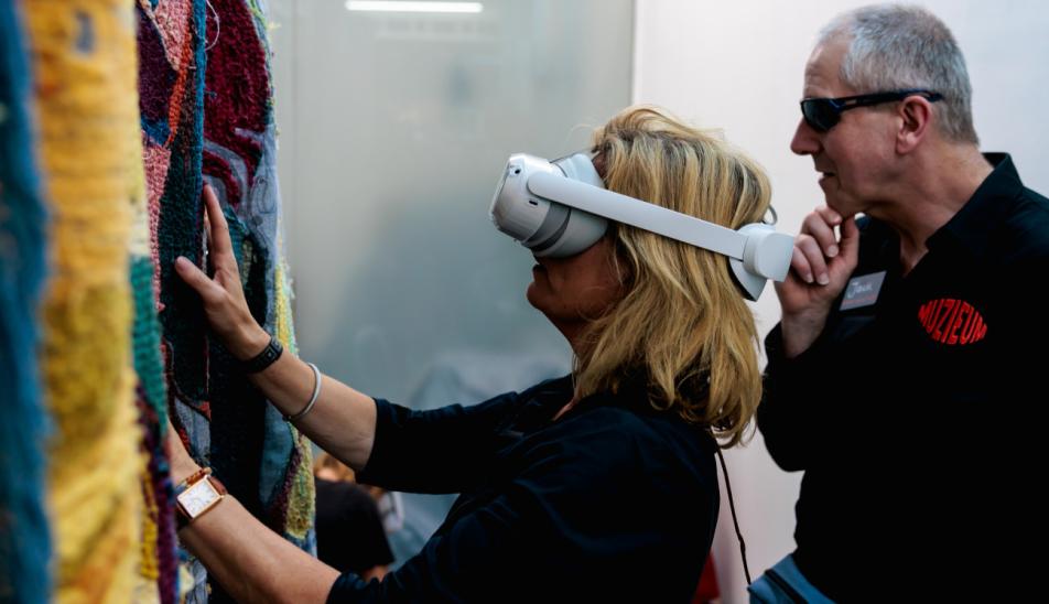 A visitor wearing an AR headset feels a colorful patchwork by Larissa Esvelt. In the background, a muSEEum guide is leading the evening.