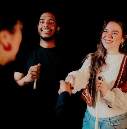 Two visitors in the Tour in absolute darkness are shaking hands with guide Melly. They are all holding a tactile cane.